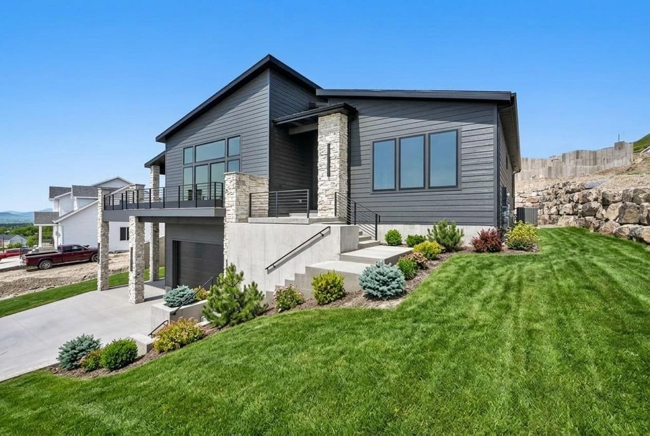 Modern two-story residential home with gray siding, stone accents, and large windows overlooking landscaped lawn and hillside setting. Built by the best home builder in Cache Valley